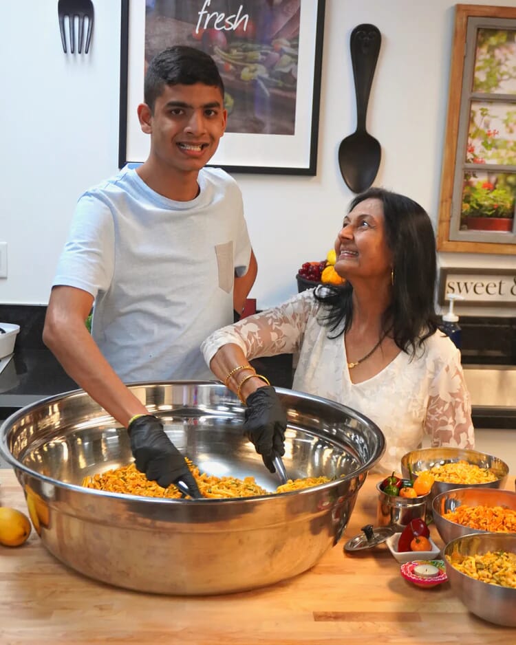 A young man and an older woman stand in a kitchen, smiling together. The man, wearing gloves, mixes food in a large metal bowl while the woman looks at him fondly. Bowls of food and ingredients for Rishi Snacks line the counter, reflecting community empowerment.