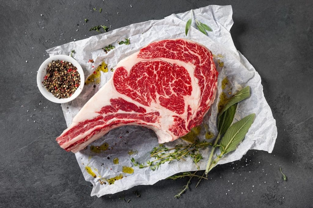 Raw bone-in ribeye steak with marbled fat sits on parchment paper, surrounded by fresh herbs and a small bowl of mixed peppercorns—a premium choice from our butcher counter’s fresh cuts.