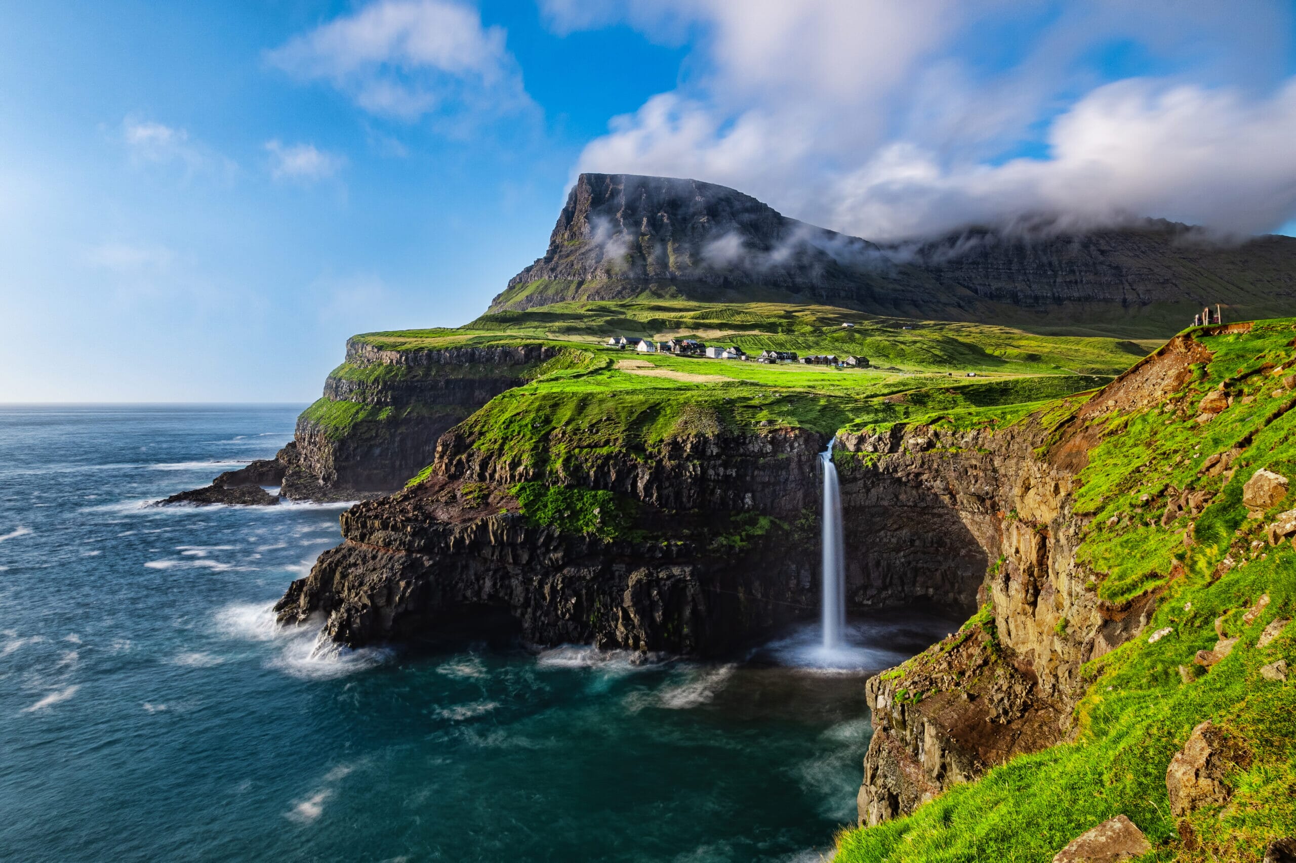 A dramatic Faroe Islands coastal landscape with steep green cliffs, a tall waterfall dropping into the ocean below, small white houses on the plateau, and a mountain partly covered in clouds under a bright blue sky.