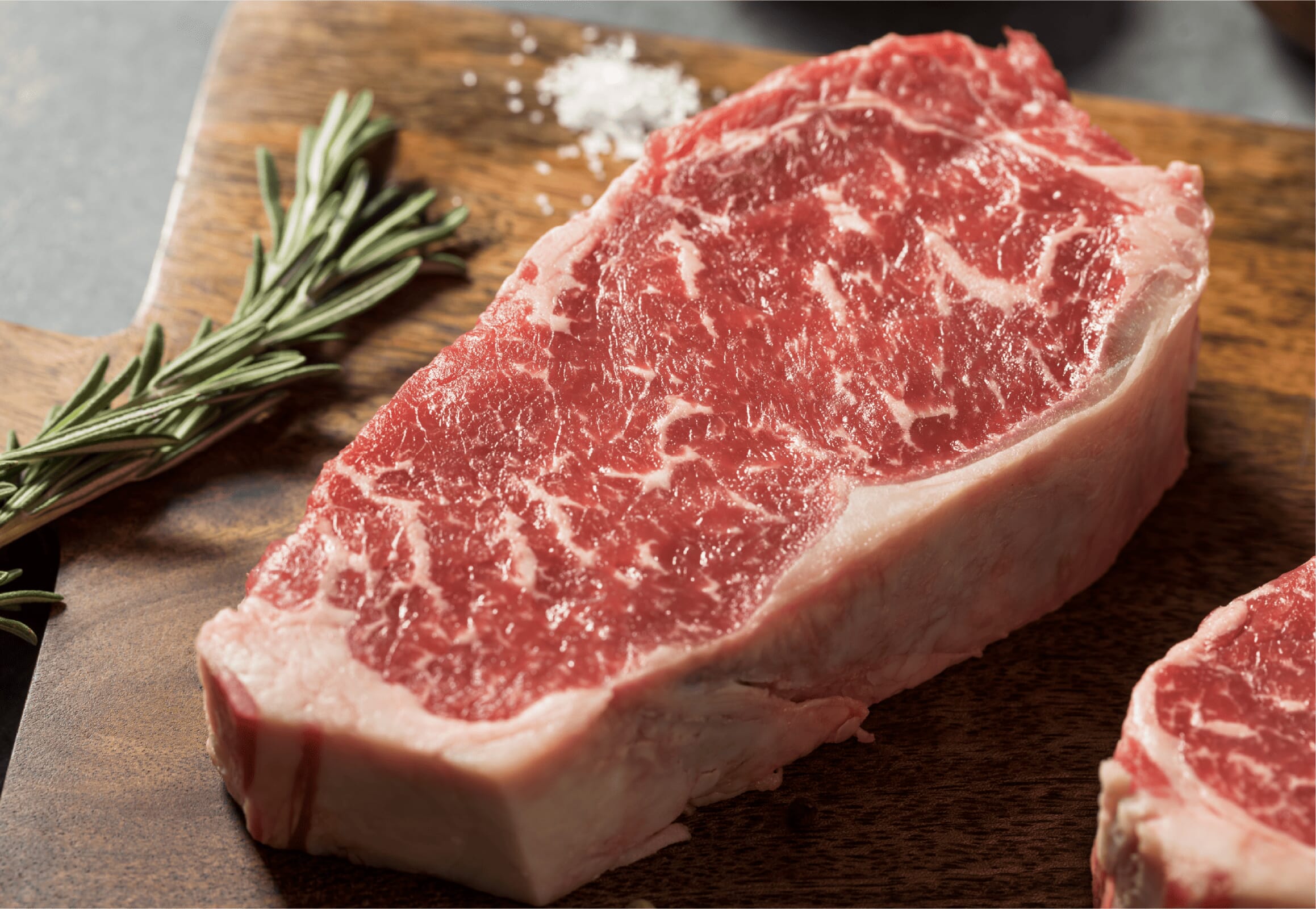 A raw, marbled steak from the butcher counter sits on a wooden cutting board next to a sprig of fresh rosemary and a small pile of coarse salt.