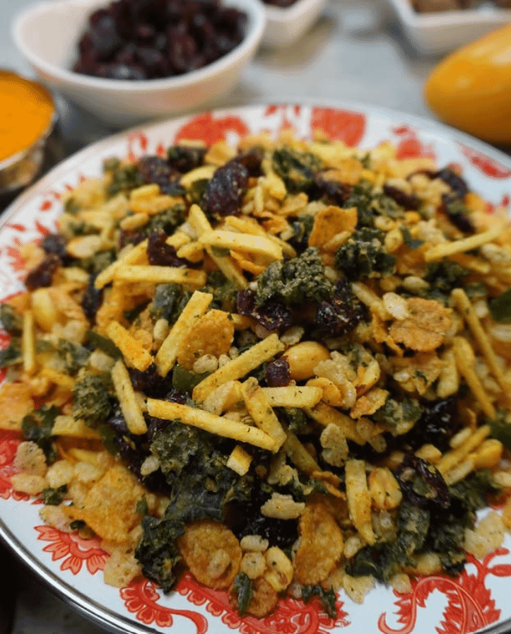 A colorful plate of Indian chivda snack mix by Rishi Snacks, featuring puffed rice, sev, dried cranberries, corn flakes, and green herbs on a floral-patterned dish. Bowls of additional ingredients highlight community empowerment in every bite.