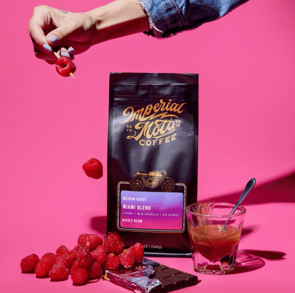 A hand sprinkles raspberries over a pile of raspberries, a chocolate bar, a glass of coffee with a spoon, and a bag of Miami's Roasters Meeria Moto Miami Blend coffee, all set against a bright pink background.