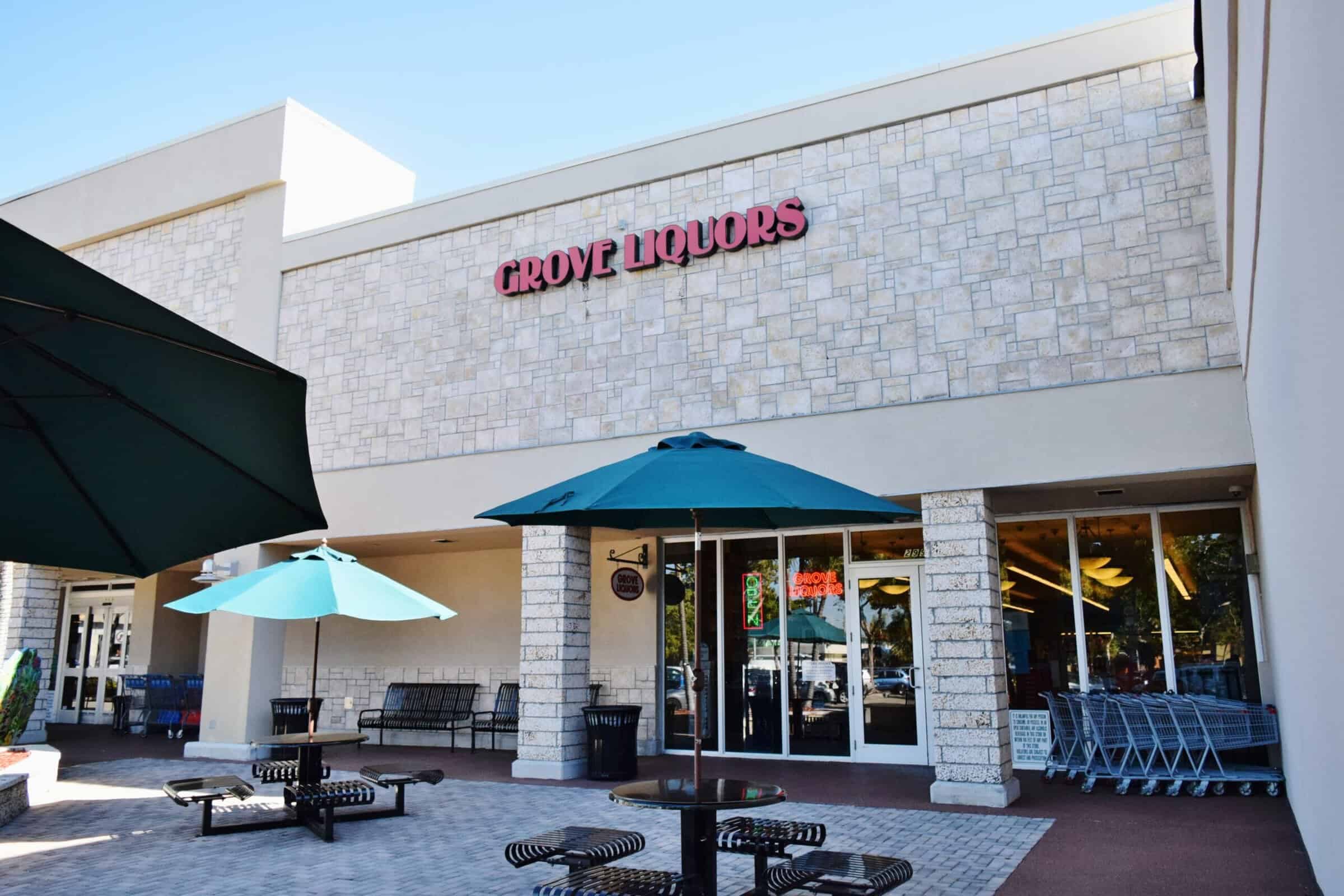 Exterior view of Grove Liquors store with stone facade, bold red Grove Liquors sign, large windows, blue and green patio umbrellas, black metal benches, and shopping carts near the entrance.