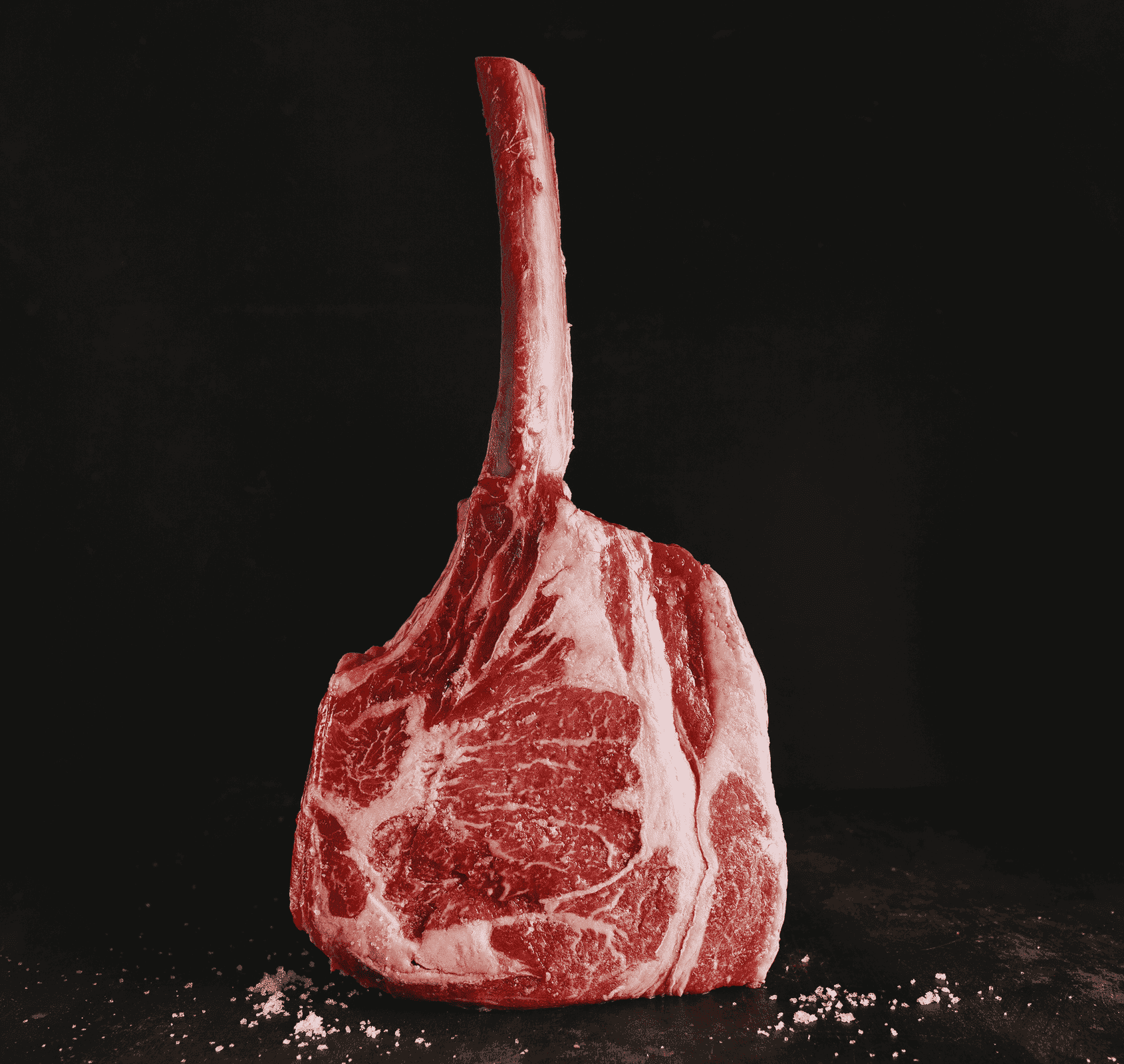 A raw tomahawk steak, showcasing premium meat selection, stands upright on a dark surface with visible marbling and a long bone. Coarse salt is sprinkled at its base, set against a striking black background.