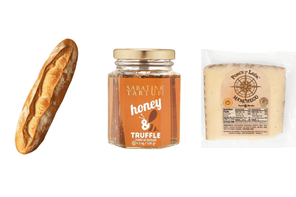 A baguette, a jar of truffle honey, and a wedge of Manchego cheese in clear packaging are arranged side by side on a white background—perfect for delicious cheese pairings.