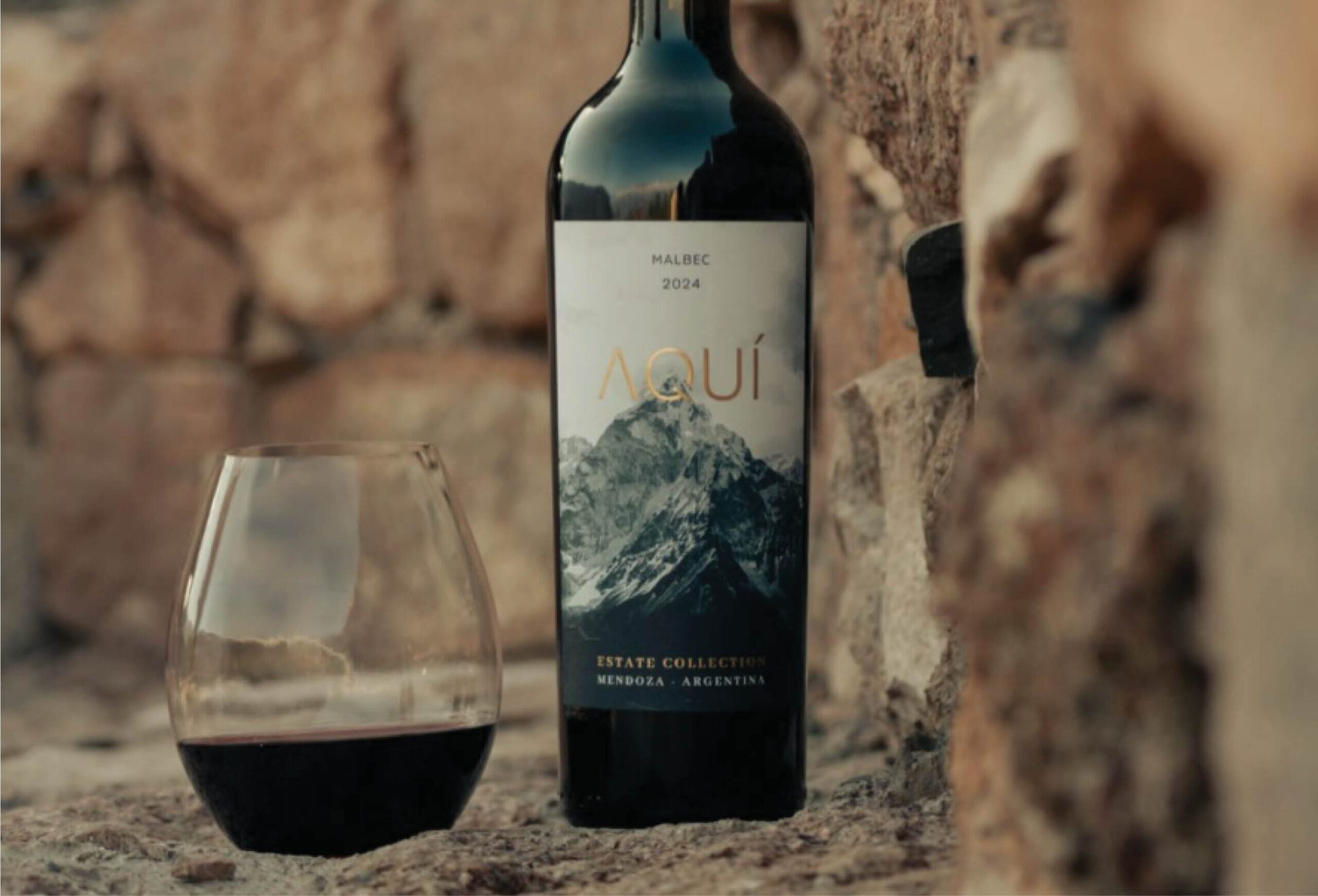 A bottle of Aquí Malbec 2024 wine and a glass of red wine rest on a stone surface, with a rustic stone wall in the background. The wine label features a mountain image and text indicating it is from Mendoza, Argentina.