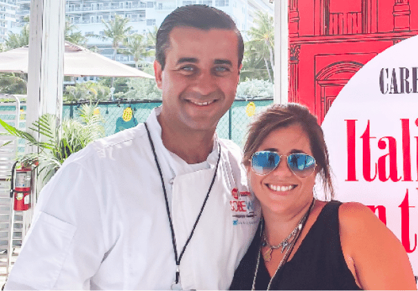 A smiling man in a chef’s coat stands next to a woman in sunglasses and a black top. Outdoors, green trees and a red sign reading Italian set the scene, hinting at delicious locally baked sweets nearby.