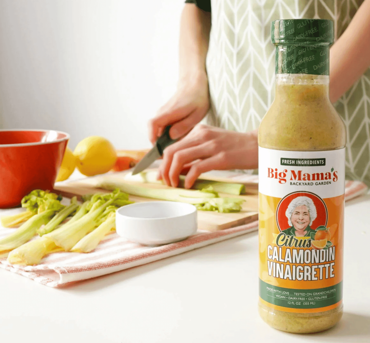 A person chops celery on a cutting board next to a red bowl, a lemon, and a striped towel. In the foreground is a bottle of Big Mama's Calamondin Vinaigrette with a green cap and colorful label.