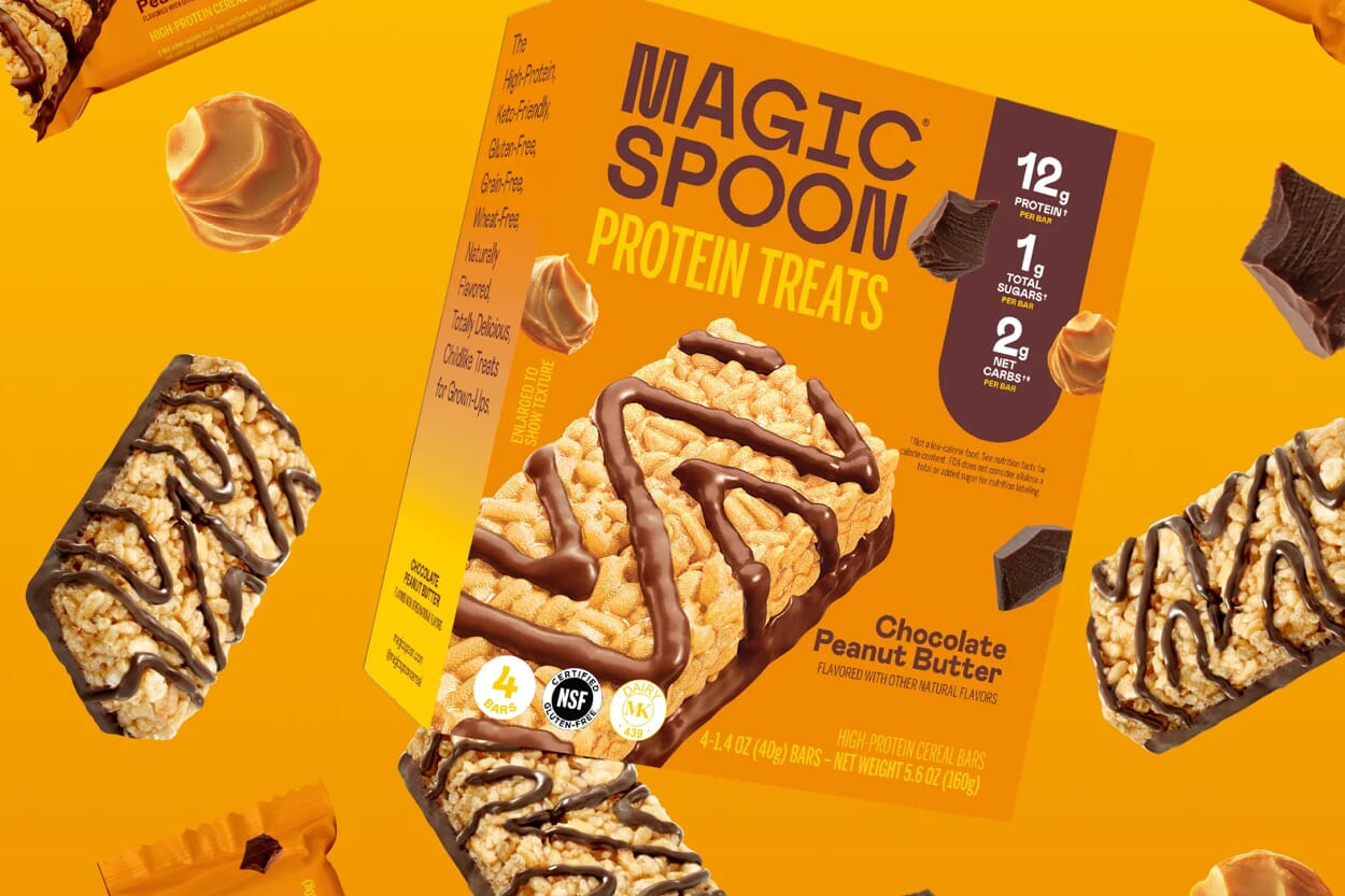 A box of Magic Spoon Protein Treats in Chocolate Peanut Butter flavor is surrounded by unwrapped bars, chocolate chunks, and peanut butter swirls on an orange background. The box highlights 12g protein, 1g sugar, and 2g net carbs per bar.