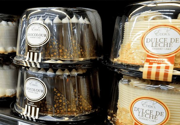 Plastic containers holding locally baked sweets like Chocolour and Dulce de Leche dessert cakes are stacked on a store shelf. The cakes feature decorative icing and toppings.