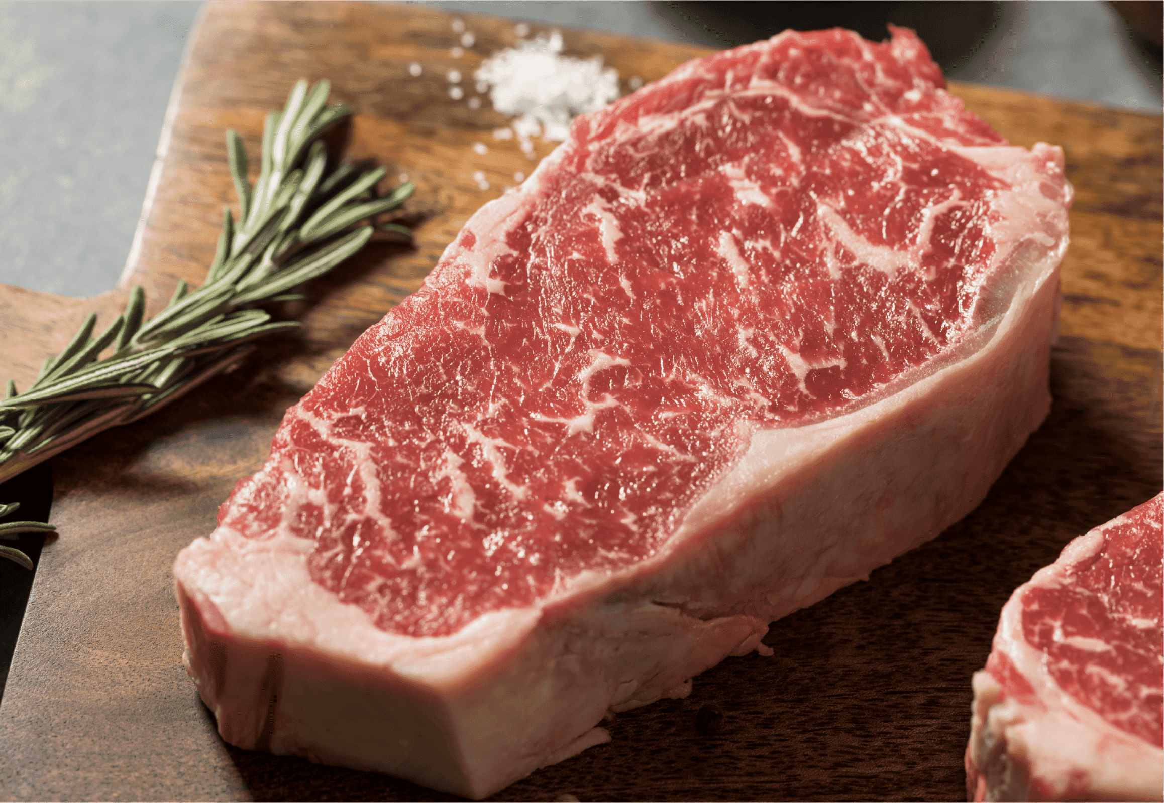 A raw, marbled steak—one of the prime beef cuts—rests on a wooden cutting board next to a sprig of rosemary and a small pile of coarse salt.