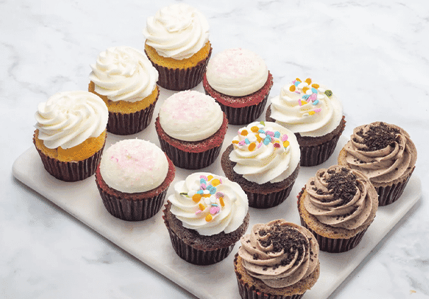 A marble slab displays twelve locally baked sweets in three rows, featuring cupcakes with various flavors and toppings like white frosting, sprinkles, pink sugar, and cookie crumbles on a white background.