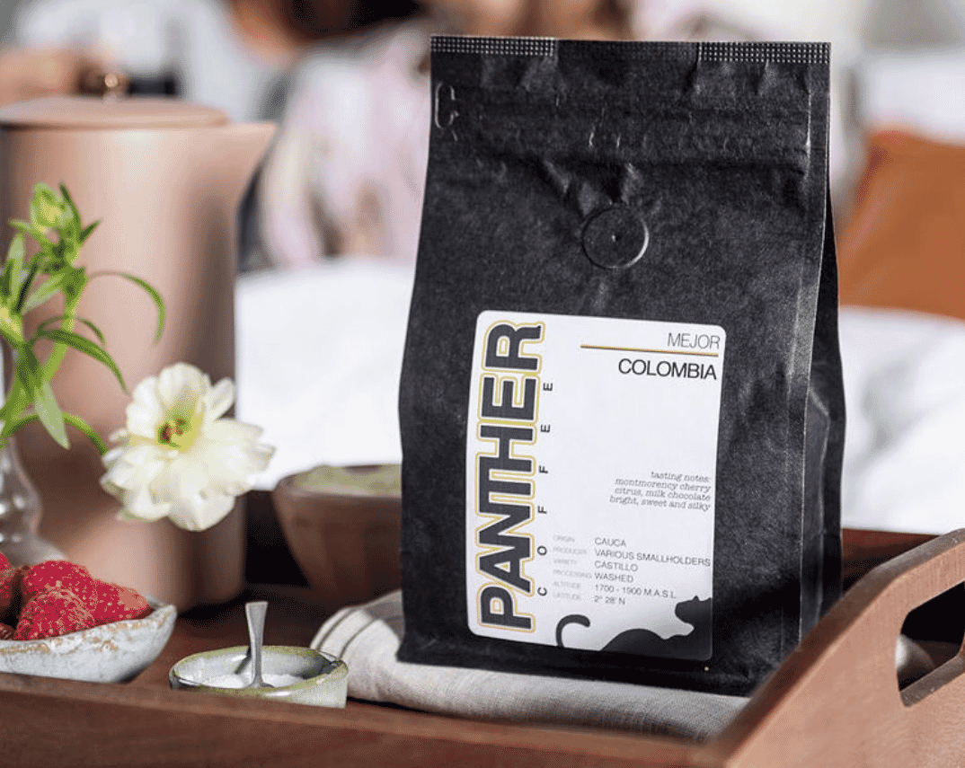 A black bag of Panther Coffee from Miami's Roasters labeled Colombia sits on a wooden tray next to a small bowl of raspberries, a flower in a vase, and a ceramic pitcher. The scene looks bright and inviting.