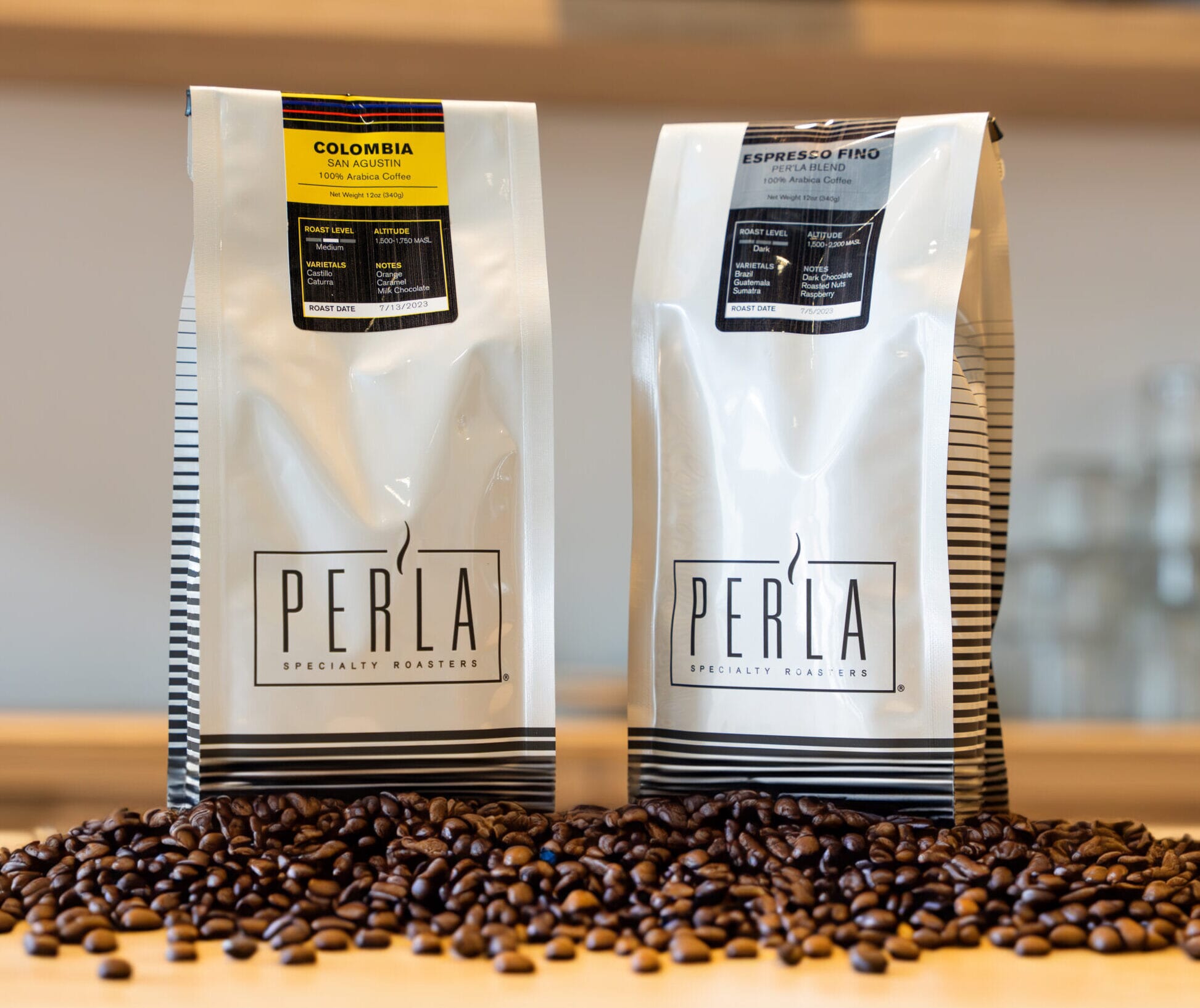 Two white Perla Specialty Roasters coffee bags stand upright on a wooden surface, surrounded by scattered coffee beans. One bag is labeled Colombia and the other Espresso Fino, reflecting the craft of Miami’s renowned roasters. Blurred shelves are in the background.