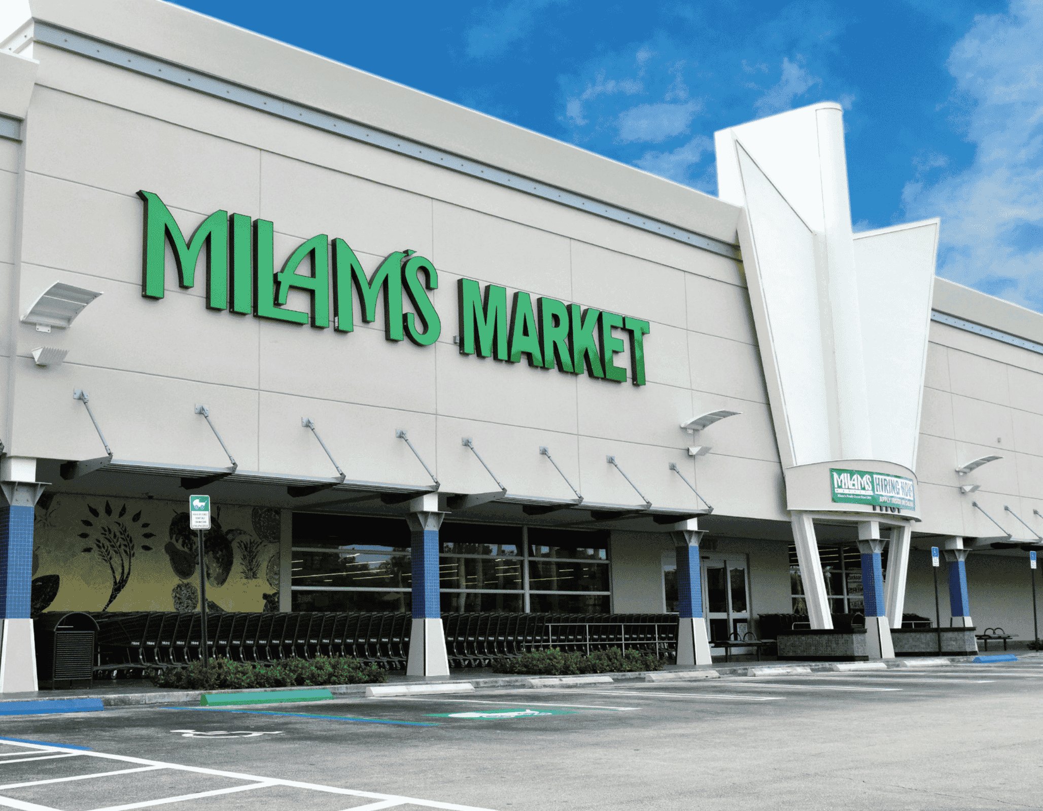 The exterior of Milams Market, a grocery store with a modern white facade and green signage, set against a blue sky with some clouds; shopping carts and empty parking spaces are visible in front.