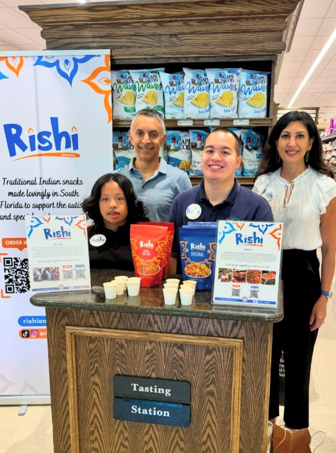 Four people stand behind a wooden tasting station with Rishi Snacks displayed. A banner and snack boxes are visible as the group smiles in a grocery store, offering samples and supporting community empowerment through healthy choices.