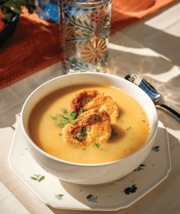 A bowl of creamy Mexican soup, inspired by a traditional recipe, topped with two pieces of toasted bread and chopped herbs, sits on a decorative plate with a glass featuring a colorful floral pattern in the background.